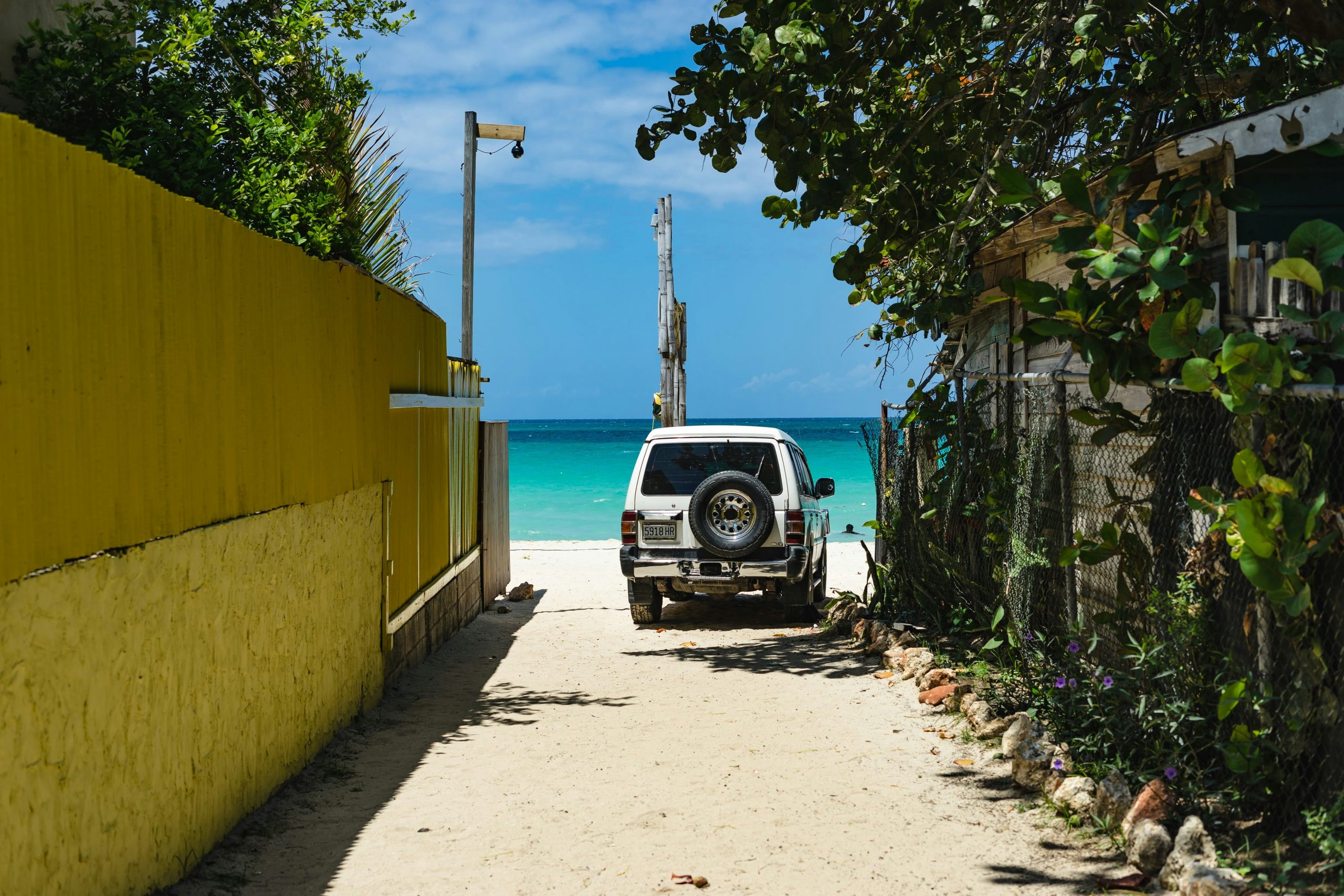 Sandy alleyway between a yellow wall and wooden fence leading to the turquoise sea in Negril Jamaica