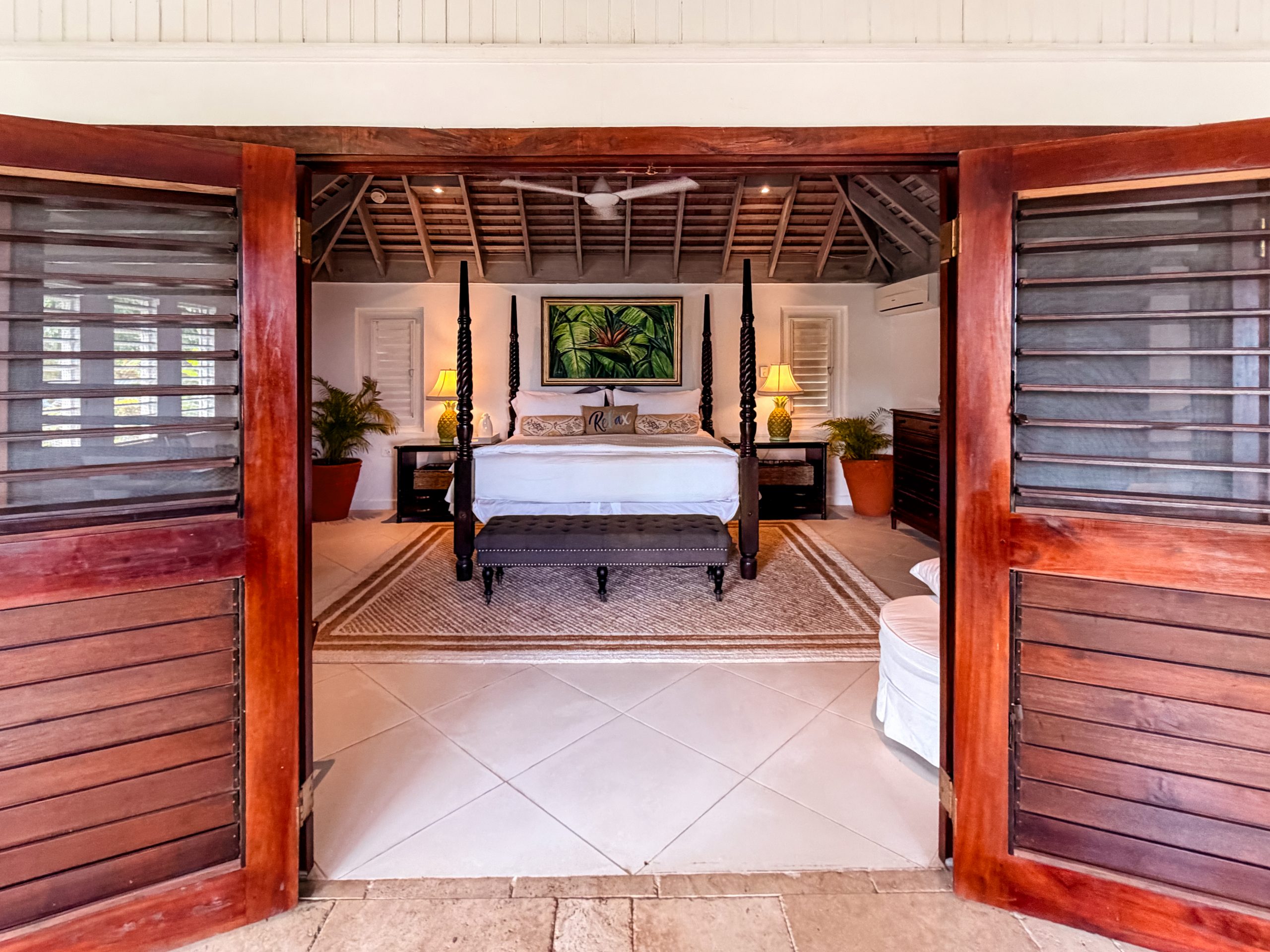 Tropical luxury villa suite with four‑poster bed, wooden doors, vaulted ceiling, and Caribbean décor at Coconut Cottage.