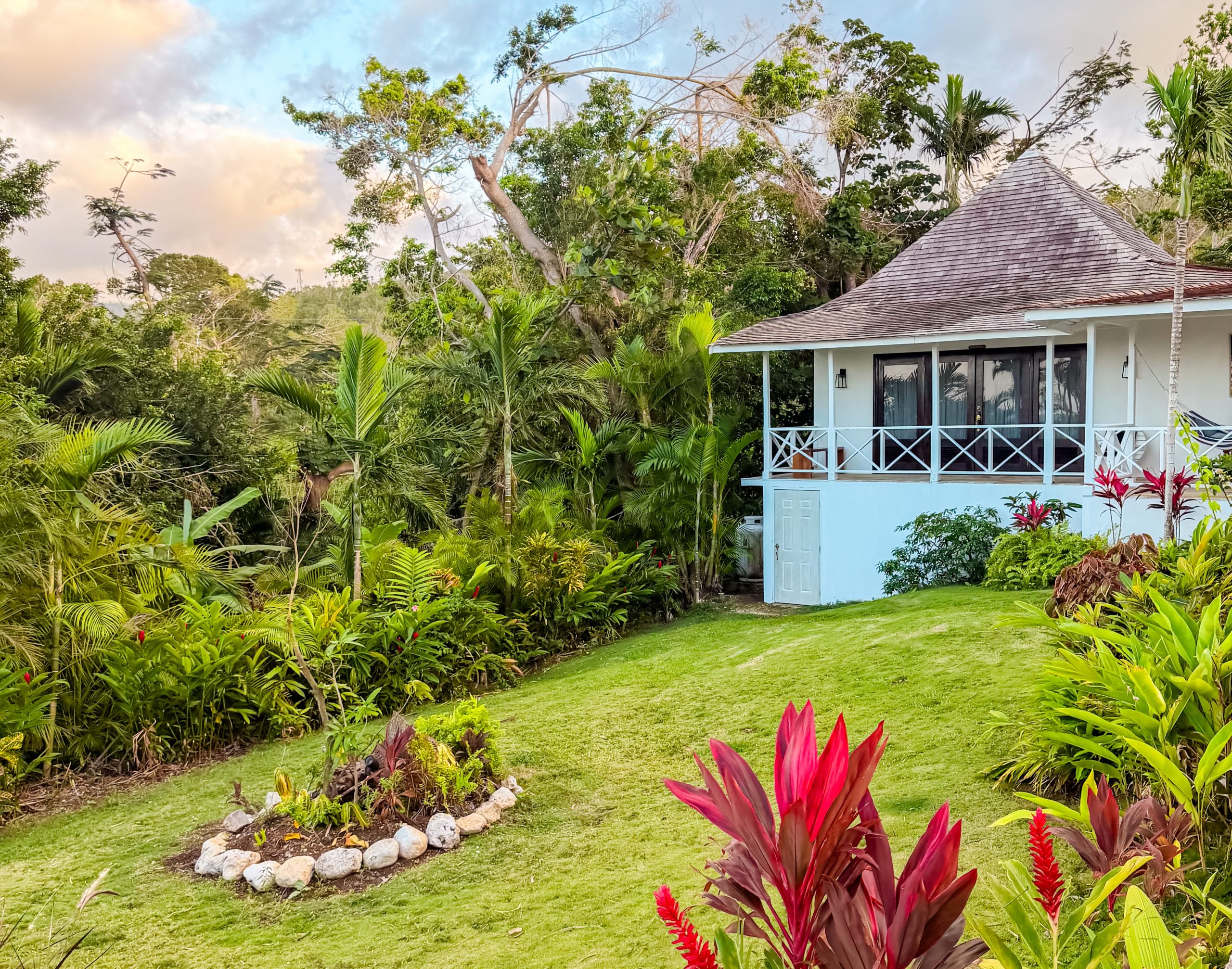 White cottage at Coconut Cottage Montego Bay surrounded by tropical plants, palm trees and a circular garden bed.
