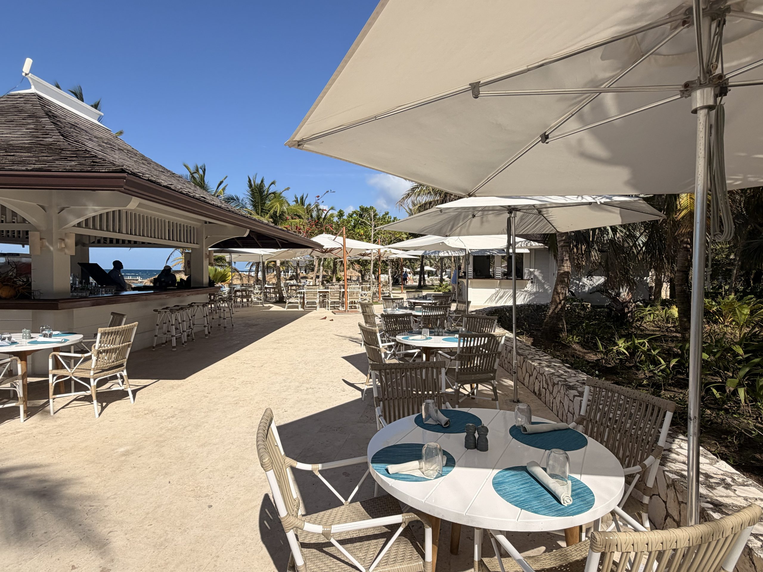 Beachside dining area at Tryall with white tables, wicker chairs and turquoise place settings under large umbrellas.