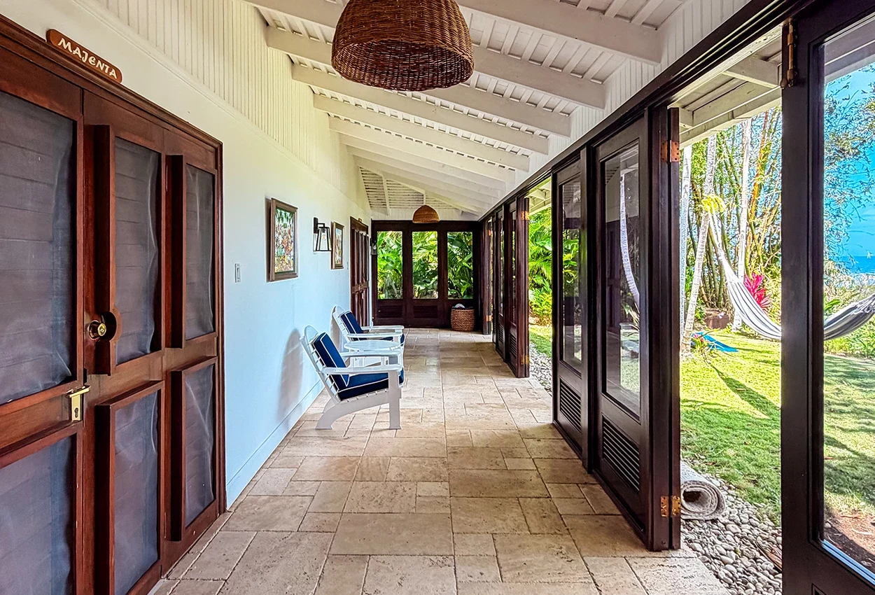 Bright verandah corridor at Coconut Cottage with white ceiling, mahogany wooden doors, blue cushioned chairs and garden views through glass doors.