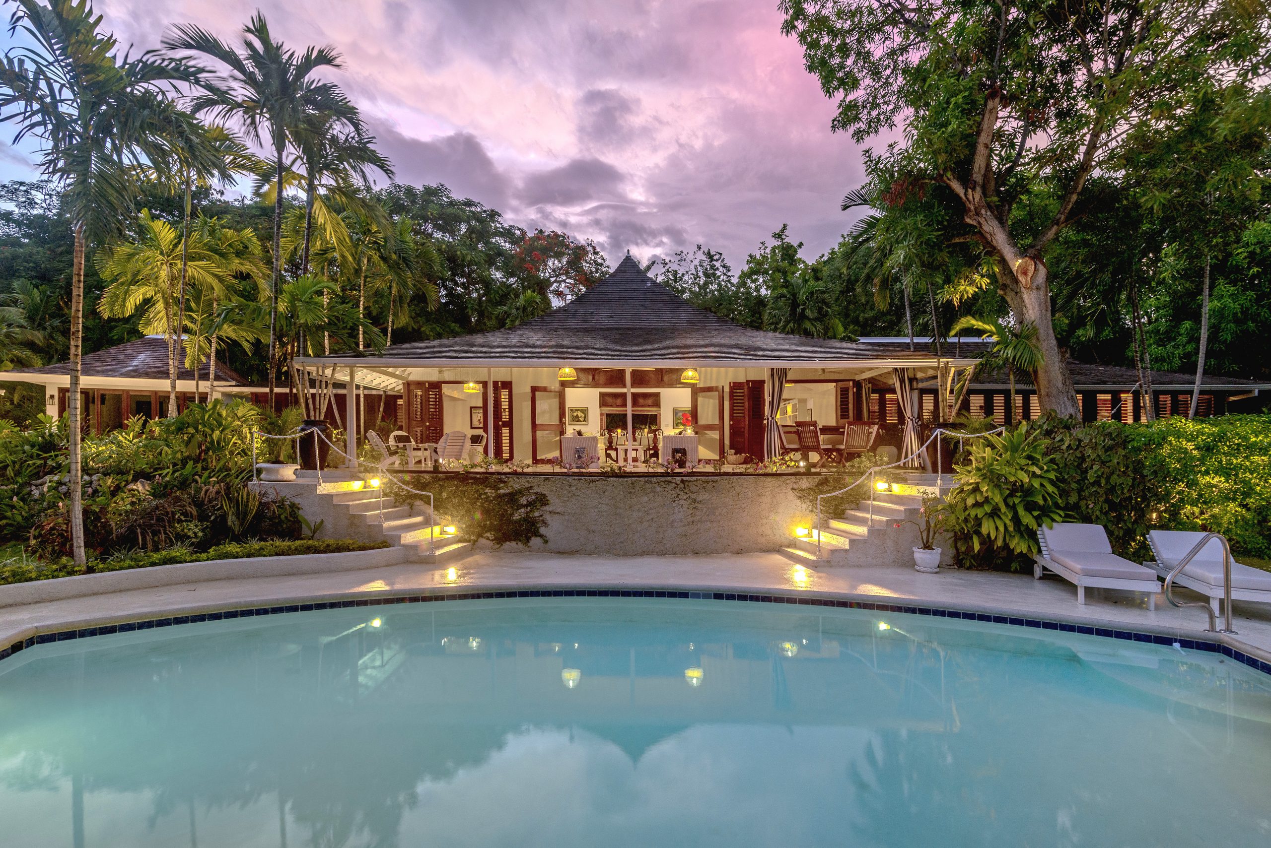 Is Montego Bay safe. Evening view of Coconut Cottage with illuminated pavilion style villa, curved pool, palm trees and sunset sky.
