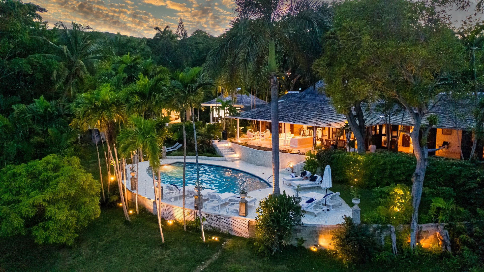 Evening view of Coconut Cottage, securely contained with illuminated pool, tropical gardens and palm trees surrounding the villa at sunset.