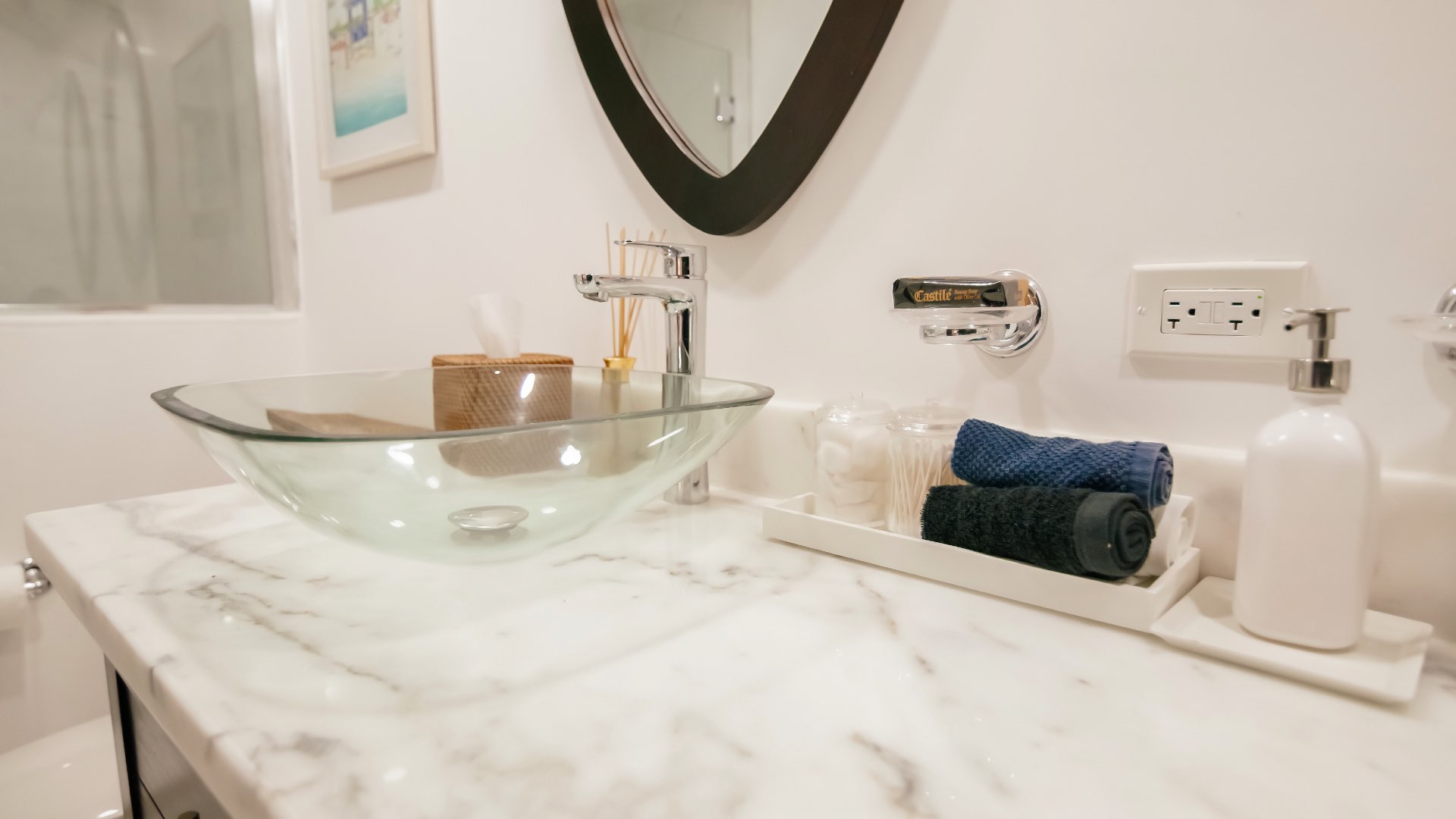 Modern Coconut Cottage bathroom with a glass vessel sink, marble countertop, rolled towels, and neatly arranged spa essentials beside a wooden‑framed mirror.