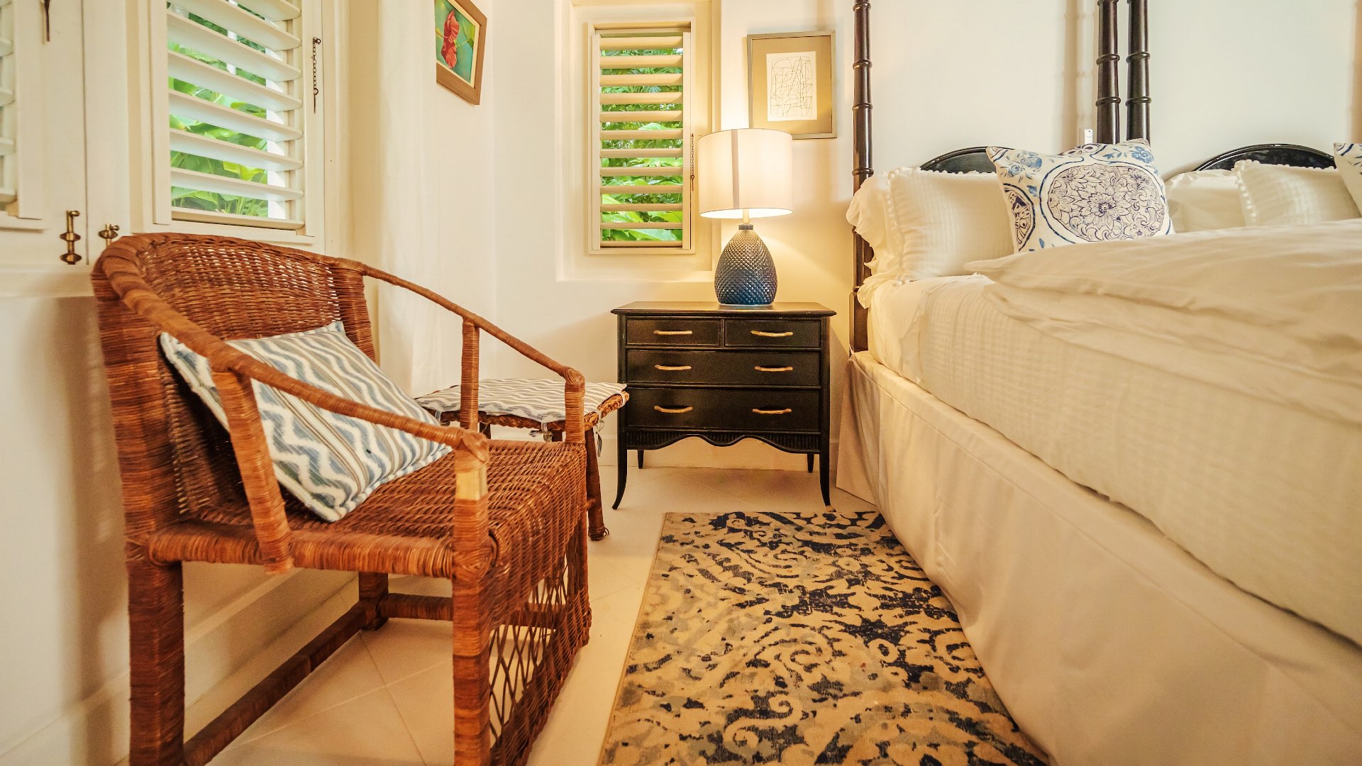 Cozy Coconut Cottage bedroom featuring a wicker chair, blue‑patterned textiles, black nightstand with a textured lamp, and a neatly made bed with white and blue decor.