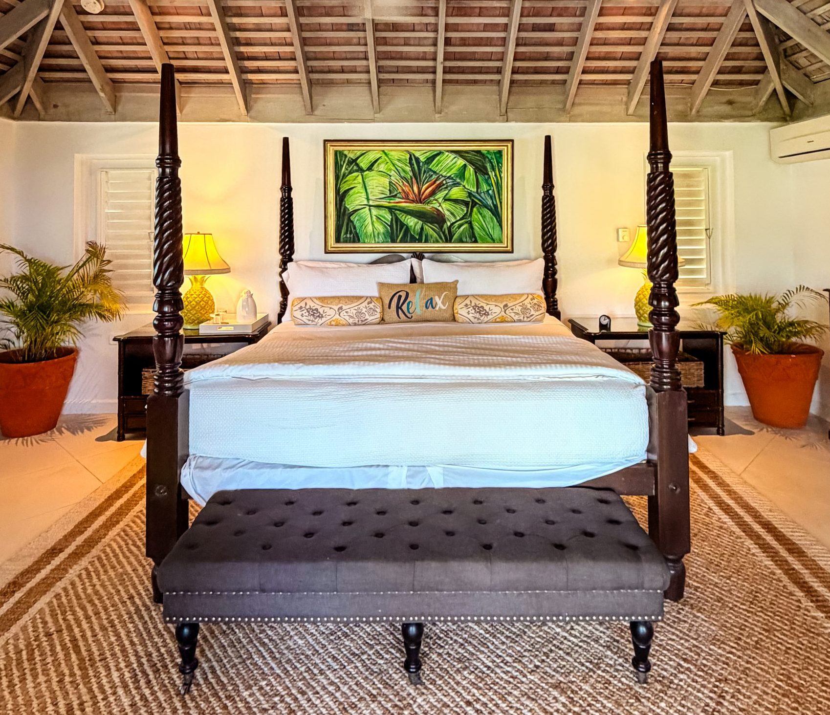 Coconut Cottage bedroom with a four‑poster bed, soft white bedding, warm ambient lighting, and a colorful tropical leaf painting that adds character without overwhelming the space.