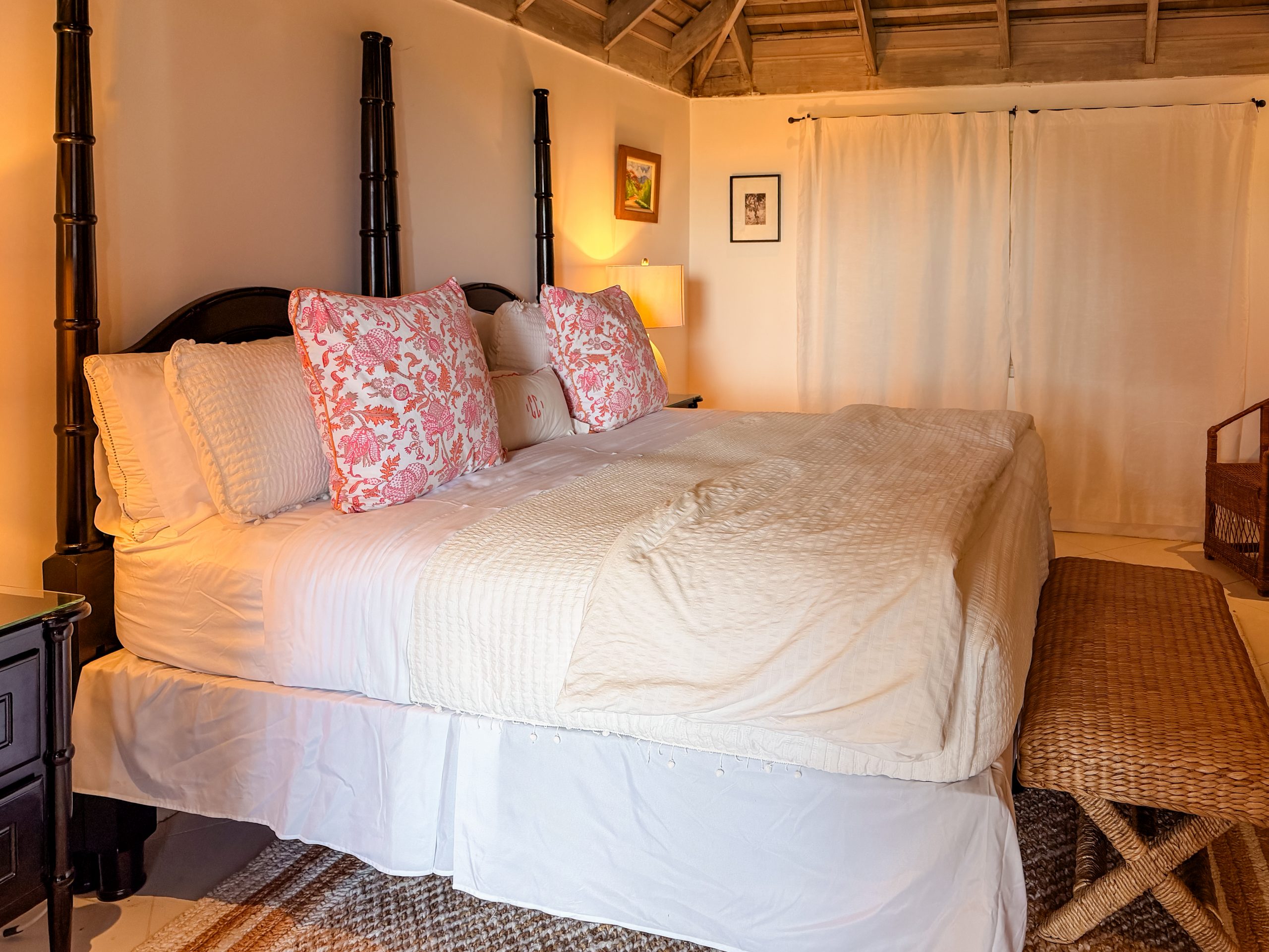 Cozy Coconut Cottage bedroom softly lit by natural daylight and warm lamp light, featuring white bedding, pink floral pillows, and wooden beams for a calm, restful atmosphere.